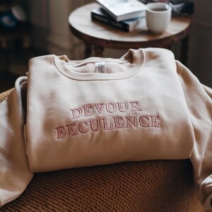 May include: A beige sweatshirt with the words "DEVOUR FECULENCE" embroidered in pink.