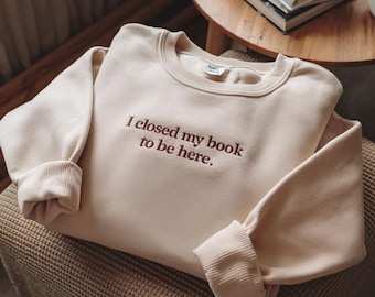 May include: A beige sweatshirt with the text "I closed my book to be here." embroidered in brown.