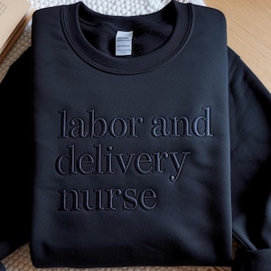 Może przedstawiać: Czarna bluza z okrągłym dekoltem z wyhaftowanym napisem "labor and delivery nurse". Bluza jest w jednolitym czarnym kolorze i wydaje się być wykonana z miękkiego materiału. Tekst jest w pasującej czarnej nici.