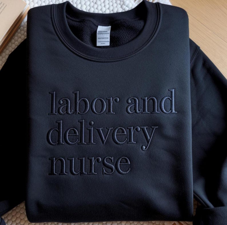 Może przedstawiać: Czarna bluza z okrągłym dekoltem z wyhaftowanym napisem "labor and delivery nurse". Bluza jest w jednolitym czarnym kolorze i wydaje się być wykonana z miękkiego materiału. Tekst jest w pasującej czarnej nici.
