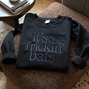 May include: Black sweatshirt with the embroidered phrase "it's frickin' bats" and bat silhouettes. The long-sleeved top is made of a soft material and is displayed on a brown patterned chair. A casual, Halloween-themed garment.