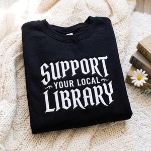 Może przedstawiać: Czarna bluza z okrągłym dekoltem i białym haftowanym napisem SUPPORT YOUR LOCAL LIBRARY wykonanym wyrazistą, stylizowaną czcionką. Napis jest wyśrodkowany na klatce piersiowej i otoczony ozdobnymi zawijasami przy słowach YOUR LOCAL.