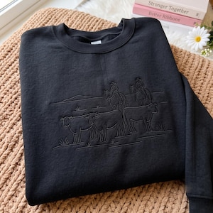 May include: A black crewneck sweatshirt featuring a tonal embroidered design of two figures on horseback herding three cattle across a landscape. The embroidery uses black thread on the black fabric, creating a subtle, textured line art illustration.