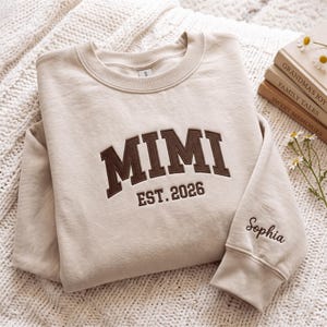 以下が含まれることがあります： 胸元にMIMI EST. 2026とダークブラウンの刺繍が施されたベージュのクルーネックトレーナー。右袖口には、ダークブラウンの筆記体でSophiaという名前が刺繍されています。