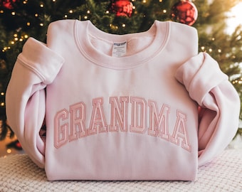 May include: A light pink crewneck sweatshirt featuring the word GRANDMA embroidered in a matching pink thread across the chest in a bold, arched collegiate-style font. The garment has long sleeves and a ribbed collar, cuffs, and hem.