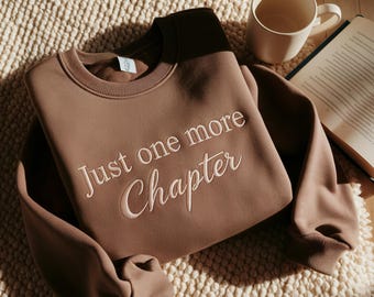 May include: A brown sweatshirt with the embroidered text "Just one more Chapter" in white. A white mug and an open book are next to the sweatshirt on a textured surface. The sweatshirt has a crew neck and long sleeves.