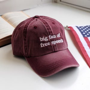 May include: A maroon baseball cap with the embroidered text "big fan of free speech" in white. The cap is made of a soft, worn material and has a curved brim. The cap is sitting on a white surface.