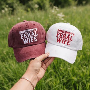 May include: Two baseball caps, one in a faded maroon color with white embroidered text and one in white with red embroidered text. Both caps feature the phrase SOMEBODY'S FERAL WIFE in a bold, decorative font across the front panels.