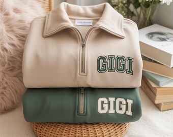 May include: Two folded sweatshirts, one beige and one green, stacked on a woven basket. Each sweatshirt has a quarter-zip collar and the name "GIGI" embroidered on the front. The beige sweatshirt has green lettering, and the green sweatshirt has white lettering.