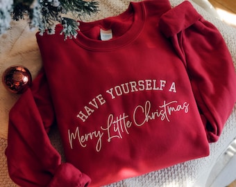 May include: A red sweatshirt with the phrase "Have Yourself a Merry Little Christmas" in white script. The sweatshirt is folded on a neutral-colored surface, with a Christmas ornament and a sprig of a Christmas tree in the background.
