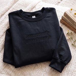 Op de afbeelding: Een zwart sweatshirt met ronde hals en de geborduurde tekst "dangerously overeducated" in bijpassend zwart garen. Het kledingstuk heeft lange mouwen met geribbelde boorden en een geribbelde halslijn, met een ton-sur-ton ontwerp op de borst.