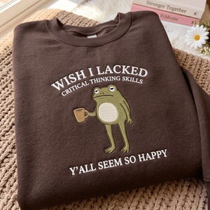 May include: A dark brown crewneck sweatshirt featuring an embroidered graphic of a green frog holding a small brown mug. The text above the frog reads WISH I LACKED CRITICAL THINKING SKILLS and the text below reads Y'ALL SEEM SO HAPPY.