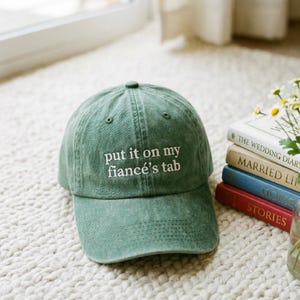 May include: A green baseball cap with the embroidered phrase "put it on my fiancé's tab" in white. The hat is made of a soft material and has a curved brim. The cap is sitting on a white, textured surface.