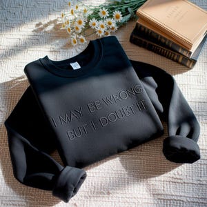 May include: Black sweatshirt with the embroidered text "I MAY BE WRONG BUT I DOUBT IT." The long-sleeved crewneck top is folded, with the sleeves rolled up. The sweatshirt is on a light-colored, textured surface, with flowers and books in the background.