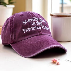 May include: A distressed purple baseball cap with the embroidered text "Morally Grey is my Favorite Color" in white. The cap has a curved brim and a worn appearance, suggesting a vintage style.