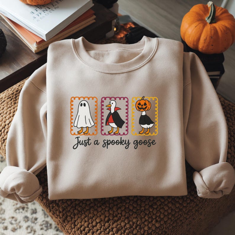 May include: A beige crewneck sweatshirt featuring a graphic of three geese in rectangular frames. The geese are dressed as a ghost, a vampire, and a pumpkin-headed figure. Below the graphic, the text reads, "Just a spooky goose" in black.