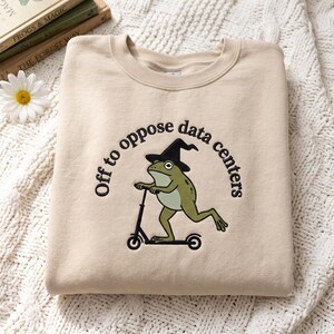Pode incluir: Uma camisola bege de gola redonda com um bordado de um sapo verde com um chapéu de bruxa preto numa trotineta preta. O texto "Off to oppose data centers" está arqueado em letras pretas por cima da ilustração do sapo.