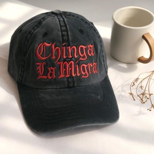 May include: Black baseball cap with the embroidered text "Chinga La Migra" in red. The cap has a curved brim and a faded, vintage look. A white mug with a wooden handle is in the background.
