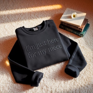 May include: Black crew neck sweatshirt with the embroidered text "I'm just here for my niece". The sweatshirt is displayed on a cream-coloured rug, with a stack of books and a flower in the background.