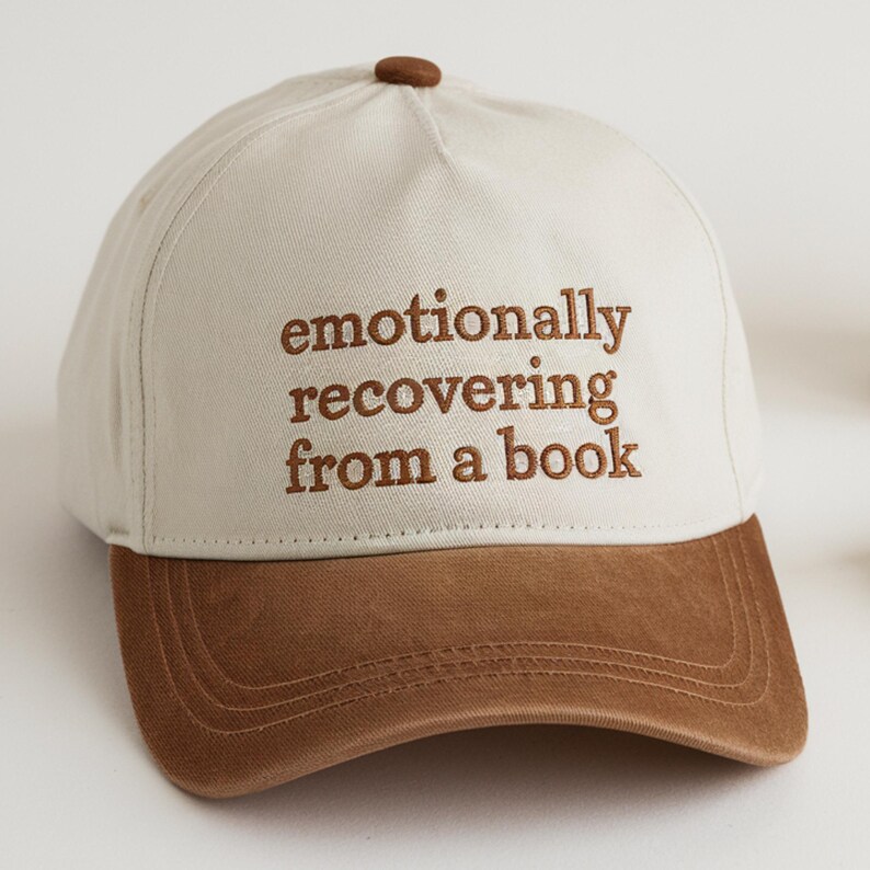 May include: A baseball cap with a cream-colored crown and a brown brim. The cap features the embroidered text "emotionally recovering from a book" in brown thread. The top button is also brown.