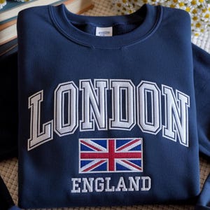 May include: Navy blue sweatshirt with the word "LONDON" in white, arched lettering. Below the word "LONDON" is a Union Jack flag and the word "ENGLAND" in white lettering. The sweatshirt is folded.