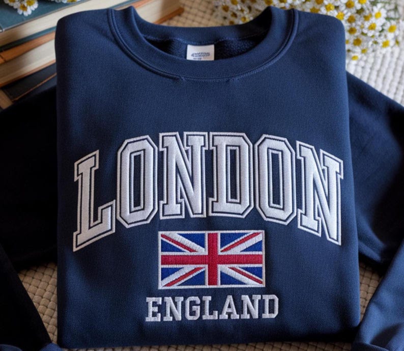 May include: Navy blue sweatshirt with the word "LONDON" in white, arched lettering. Below the word "LONDON" is a Union Jack flag and the word "ENGLAND" in white lettering. The sweatshirt is folded.