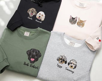 May include: Five sweatshirts with embroidered pet portraits. A black hoodie with two dogs, a pink sweatshirt with two cats, a green sweatshirt with a black dog, a gray sweatshirt with two dogs, and a pink sweatshirt with a red heart on the sleeve.