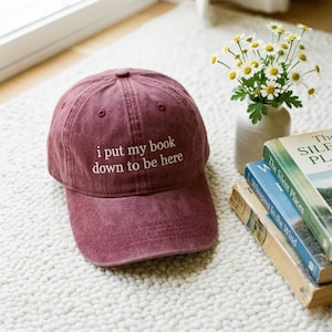 Può includere: Cappellino da baseball bordeaux con la scritta bianca "i put my book down to be here". Il cappellino è su un tappeto bianco accanto a un vaso di fiori e una pila di libri.