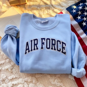 May include: A pale blue sweatshirt featuring the words "AIR FORCE" in navy blue lettering. The garment is neatly folded, with the sleeves tucked in. An American flag is partially visible in the background.