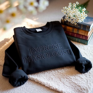 May include: A black crewneck sweatshirt with the embroidered text "EXERCISE PHYSIOLOGIST". The sweatshirt is folded on a soft, light-colored surface, with a small bouquet of white flowers and a stack of books in the background.