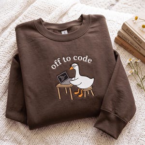 May include: A dark brown crewneck sweatshirt featuring an embroidered graphic of a white goose wearing glasses, sitting at a small desk with a laptop. The text "off to code" is embroidered in white above the goose, which has orange feet and beak.
