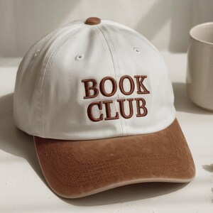 Pu&ograve; includere: Cappellino da baseball bianco con visiera e bottone marrone. Le parole "BOOK CLUB" sono ricamate in marrone sul davanti. Un accessorio casual.