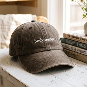 Puede incluir: Una gorra de béisbol de color marrón oscuro, teñida con pigmento, con visera curva y un botón en la parte superior. La parte frontal de la gorra presenta el texto "body builder" bordado en letras minúsculas blancas sin remates.