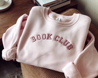 Puede incluir: Una sudadera rosa con las palabras "BOOK CLUB" bordadas en un color rosa más oscuro.