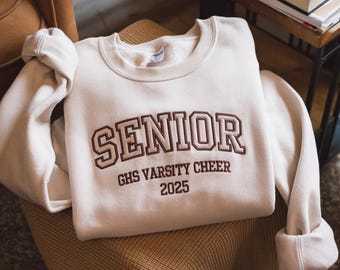 Puede incluir: Sudadera color crema con la palabra "SENIOR" en letras universitarias marrones. Debajo de la palabra "SENIOR" está "GHS VARSITY CHEER 2025". La sudadera está doblada sobre una silla.