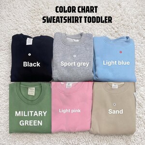 May include: A color chart for toddler sweatshirts featuring six crewneck pullovers in black, sport grey, light blue, military green, light pink, and sand. The text reads COLOR CHART SWEATSHIRT TODDLER, with labels identifying each color.