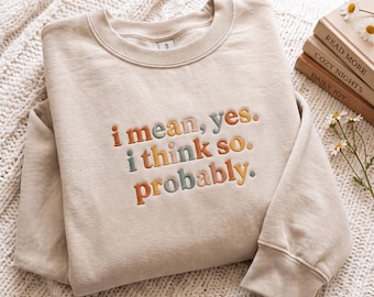 May include: A beige crewneck sweatshirt featuring embroidered text in shades of orange, teal, and brown. The text reads: i mean, yes. i think so. probably. The sweatshirt has a ribbed collar, cuffs, and hem, with a soft, solid-colored fabric construction.