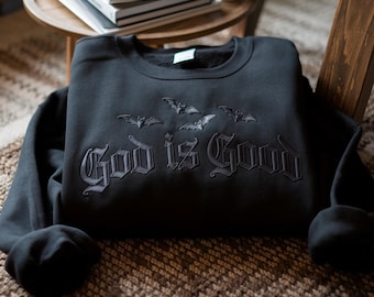 Könnte beinhalten: Ein schwarzes Sweatshirt mit Rundhalsausschnitt und einer tonalen schwarzen Stickerei von drei fliegenden Fledermäusen über dem Text "God is Good" in einer gotischen Schriftart. Das Kleidungsstück hat lange Ärmel und einen gerippten Ausschnitt.