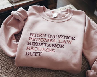 Puede incluir: Una sudadera de cuello redondo en color rosa claro con texto bordado en rojo oscuro que dice: WHEN INJUSTICE BECOMES LAW RESISTANCE BECOMES DUTY. La prenda tiene mangas largas con puños acanalados y un cuello acanalado de diseño sencillo.