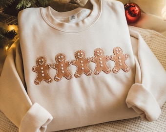 May include: A cream-colored sweatshirt featuring an embroidered design of five gingerbread men with smiling faces. The gingerbread men are brown with white icing details. The sweatshirt is folded on a white knitted blanket, with a Christmas tree and ornament in the background.