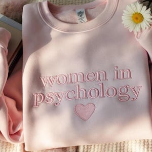 Może przedstawiać: Jasnor&oacute;żowa bluza z haftowanym napisem "women in psychology" i sercem. Biała stokrotka z ż&oacute;łtym środkiem umieszczona w pobliżu rękawa. Bluza wykonana jest z miękkiego materiału.
