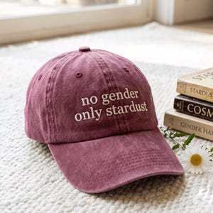 May include: A maroon baseball cap with a six-panel construction and a curved brim. The front of the cap features the embroidered white text "no gender only stardust" in a clean, sans-serif font. The fabric has a slightly faded, textured finish.