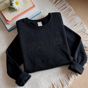 May include: Black crewneck sweatshirt with the phrase "CARL'S A MESS" embroidered across the chest. The sweatshirt is folded on a white rug with a stack of books and a daisy in the background.