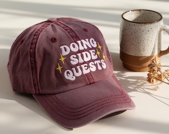 May include: A maroon baseball cap with the words "DOING SIDE QUESTS" embroidered in white with yellow star accents. A ceramic mug with a brown base and speckled white top sits in the background.