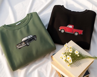 May include: Two crew neck sweatshirts, one olive green and one black, each with a car graphic. The olive green sweatshirt features a black car, while the black sweatshirt displays a red pickup truck. A bouquet of daisies rests on a stack of books.