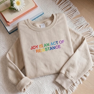 以下が含まれることがあります： 虹色の「JOY IS AN ACT OF RESISTANCE」の刺繍が施されたベージュのスウェットシャツ。スウェットシャツは、クルーネックとリブ編みの袖口が付いています。柔らかい素材で作られており、白いラグの上に置かれています。