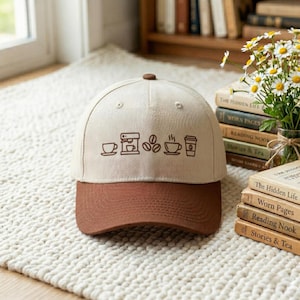 Puede incluir: Gorra de béisbol beige y marrón con un diseño temático de café. La gorra presenta una visera marrón y una corona beige con un diseño de taza de café, cafetera, granos y vaso para llevar. La gorra está sobre una alfombra blanca.