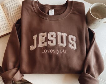 May include: A brown sweatshirt with the text "JESUS loves you" embroidered in a light beige color. The sweatshirt is folded and placed on a light-colored surface, with a book and a mug visible in the background.