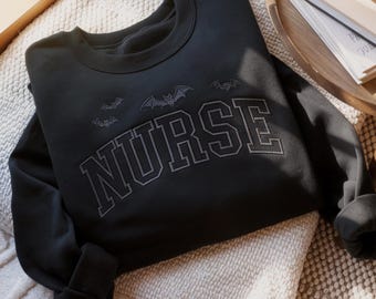 May include: Black crewneck sweatshirt with the word "NURSE" in a collegiate font, and three bat designs above the text. The sweatshirt is folded on a cream-colored blanket, with a wooden tray and a stack of books in the background.