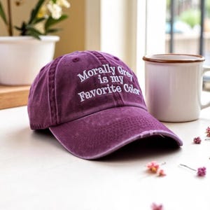 May include: A distressed purple baseball cap with the embroidered text "Morally Grey is my Favorite Color" in white. The cap is sitting on a white surface next to a white mug with a brown rim.